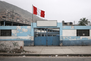 Fachada de colegio con paredes celestes y blancas descascaradas, rejas azules, bandera peruana, acera con basura y un cerro con casas al fondo.