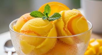 Cuenco de cristal con varias bolas de sorbete de mango y naranja, adornado con menta y una rodaja de naranja. El recipiente presenta condensación.