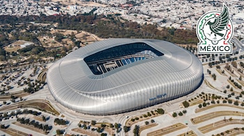 Vista aérea de un estadio moderno rodeado de estacionamientos y vegetación, con un escudo futbolístico arriba a la derecha.