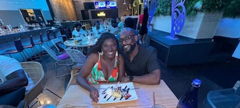 Nancy Metayer y Vince Staples sonríen a la cámara mientras están sentados en una mesa con un postre y bebidas en un restaurante