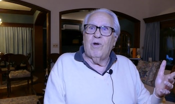 Primer plano de un hombre mayor con gafas y cabello blanco que gesticula con una mano en una sala de estar