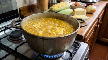 Olla metálica de locro de choclo hirviendo en una estufa de gas. Se ven choclos frescos, queso, cebolla y huevos en el mostrador de madera al fondo.