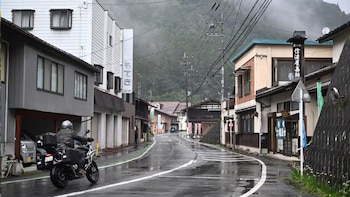 Una persona en motocicleta transita por una calle mojada con edificios a los lados y montañas cubiertas de niebla al fondo