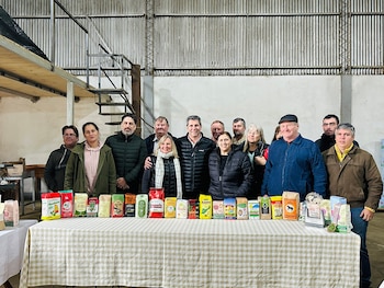 Un grupo de trece personas, incluyendo a Sergio Uñac, posa detrás de una mesa con paquetes variados de yerba mate. Fondo de pared y escaleras metálicas