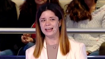 Primer plano de una mujer joven con cabello castaño y mechas claras, vistiendo un blazer color crema, un micrófono pegado a la mejilla y una sonrisa expresiva