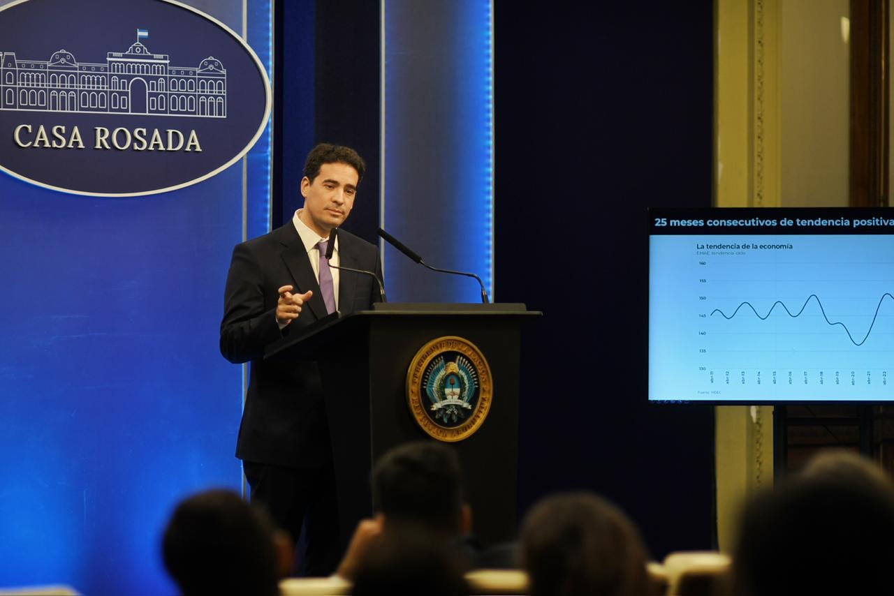 Adrián Ravier, el nuevo vocero presidencial, se dirige a la prensa en la Casa Rosada mientras un gráfico sobre la tendencia económica del país se proyecta en una pantalla