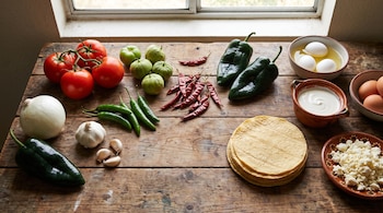 Jitomates, tomates verdes, chiles, cebolla, ajo, tortillas de maíz, huevos, crema y queso fresco acomodados sobre una mesa de madera.