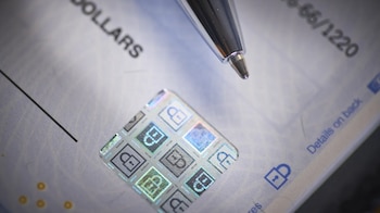 Primer plano de un cheque con un bolígrafo encima, mostrando un holograma de seguridad con símbolos de candado y texto parcial como 'DOLLARS'