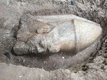 Fragmento de estatua de piedra esculpida con forma de cabeza con corona, tendido sobre la tierra en una zanja de excavación