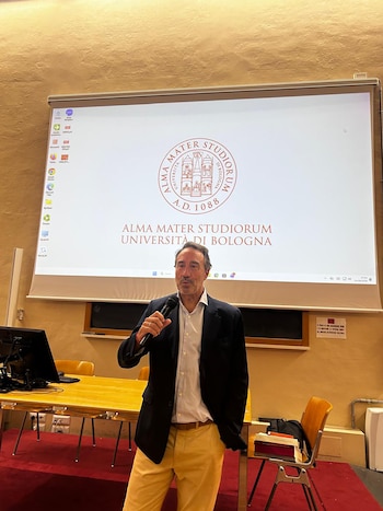 Mariano Llorens, de pie y sosteniendo un micrófono, habla frente a una pantalla de proyección con el logo de la Universidad de Bolonia en un aula