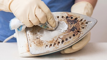Manos con guantes amarillos aplican pasta blanca con un paño sobre la base metálica con manchas oscuras de una plancha.