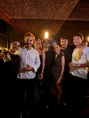Grupo de siete amigos sonriendo y posando para la cámara en un espacio interior con iluminación cálida, con algunos sosteniendo bebidas
