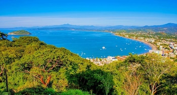 Vista aérea de una bahía azul con barcos, una isla verde, una playa con edificios y vegetación densa, y montañas al fondo bajo cielo azul