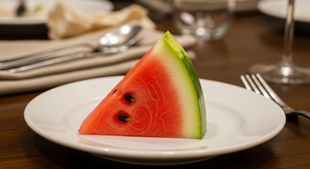 Una rodaja triangular de sandía roja con semillas negras y corteza verde y blanca, servida en un plato blanco sobre una mesa de madera oscura. Cubiertos y vasos borrosos al fondo.