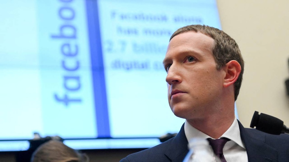 Primer plano de Mark Zuckerberg, vestido con traje oscuro y corbata morada, mirando hacia la derecha, con una pantalla borrosa que muestra 'facebo' en el fondo