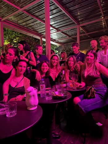 Grupo de personas en un evento nocturno con iluminación magenta. Varias mujeres y hombres sonríen y posan en un ambiente festivo con mesas y bebidas