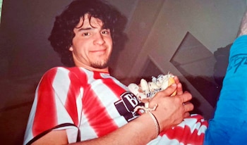 Hombre joven con cabello oscuro rizado y barba, sonriendo, viste una camiseta de rayas rojas y blancas con logo. Sostiene un objeto pequeño y claro