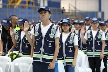 Grupo de jóvenes de pie, algunos con la mano derecha levantada, visten chalecos azules reflectantes y gorras azules. Fondo de un estadio
