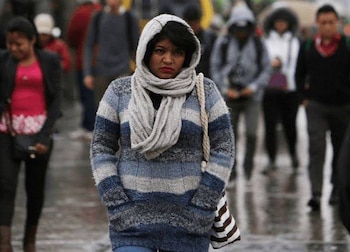 Personas con abrigos, bufandas, gorros y paraguas caminan por una calle húmeda