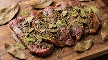 Varias piezas de carne cruda cubiertas con hojas secas de aguacate enteras y picadas, sobre una tabla de madera con hojas de aguacate secas esparcidas.