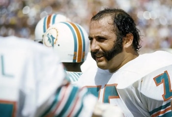 Primer plano del exjugador Manny Fernandez sonriendo en su uniforme blanco de los Miami Dolphins con el número 75 y bigote prominente en el campo