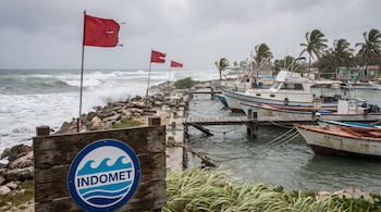 Cartel de INDOMET en primer plano, con olas altas y banderas rojas en una costa caribeña rocosa. Barcos amarrados en un puerto con cielo nublado y viento.