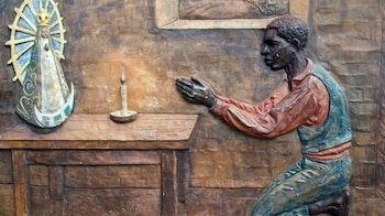 Bajorrelieve con hombre arrodillado de perfil, manos unidas. Estatua de la Virgen de Luján y vela encendida sobre mueble de madera
