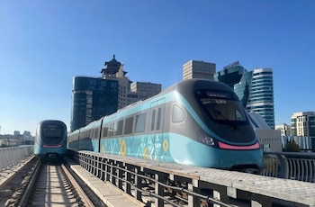 Dos trenes modernos, azules y grises, del Astana LRT en una vía elevada con un horizonte urbano y cielo azul claro de fondo