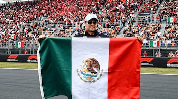 Un hombre con gorra y gafas oscuras sostiene la bandera de México desplegada en una pista frente a gradas con público.
