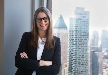 Mujer con cabello largo, gafas, camisa blanca y blazer negro con brazos cruzados. Se para junto a una ventana con un collar de dije, y un horizonte urbano borroso
