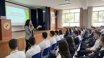 Una mujer con micrófono habla a un grupo de estudiantes. Una pantalla proyecta el texto "Salud Mental y Bienestar Escolar". Hay un atril con un logo escolar.