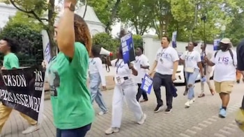 Personas diversas marchan en la calle, con carteles que dicen "Protect Our Vote" y pancartas, una con "Jackson". Algunos visten camisetas con "MS VOTES"