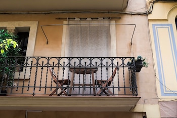 Balcón con barandilla de hierro forjado, dos sillas de madera plegables, una mesa de madera cuadrada, una planta, una ventana con cortina y cables eléctricos en la fachada