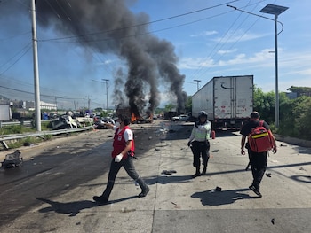 Varios socorristas con uniformes blancos y rojos inspeccionan un automóvil negro destruido junto a la estructura de un camión