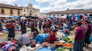 Mercado concurrido en Guatemala: Se observan numerosos vendedores y compradores entre puestos de frutas, verduras, textiles y comida en la calle, con edificios coloniales de fondo.