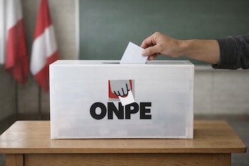 Mano insertando una cédula en urna electoral blanca con logo de ONPE en mesa de madera clara. Fondo con pizarrón verde y banderas peruanas desenfocadas.