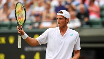 Primer plano de Axel Geller, un hombre joven con gorra blanca y polo blanco, sosteniendo una raqueta de tenis negra y amarilla, mirando hacia un lado en una cancha