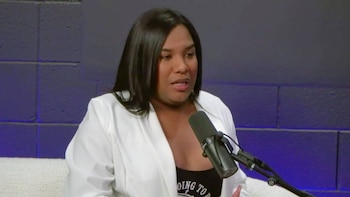Primer plano de una mujer con cabello oscuro, vistiendo una chaqueta blanca y una camiseta negra, hablando a un micrófono de estudio