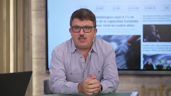 Hombre de mediana edad con bigote y gafas, camisa azul claro, hablando y gesticulando con las manos frente a una pantalla grande con texto y gráficos