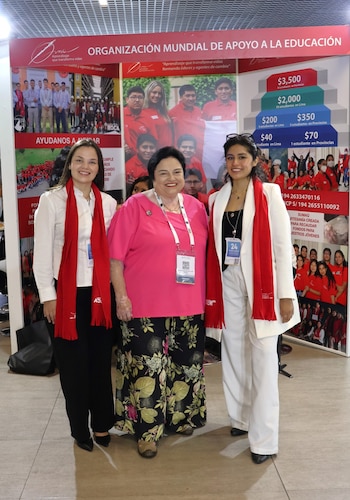 Tres mujeres de pie posan ante un panel publicitario rojo y blanco de la Organización Mundial de Apoyo a la Educación