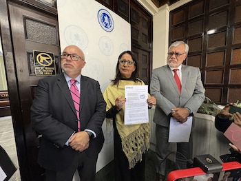 Tres expertos, dos hombres y una mujer, posan en una sala. La mujer central, con un chal amarillo, sostiene un documento. Detrás, una puerta de madera y un cartel de prohibición de armas