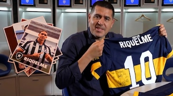 Juan Román Riquelme, hombre de mediana edad, sostiene una camiseta azul y oro de Boca Juniors con el número 10. A su izquierda, se ven fotos de un Riquelme joven en otras camisetas.