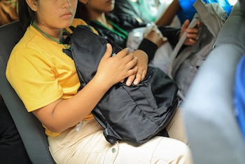 Joven sentada de perfil, con camiseta amarilla y pantalón beige, sosteniendo una mochila negra en su regazo. Sus manos reposan sobre la bolsa