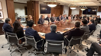 Un grupo de aproximadamente veinte personas se sienta alrededor de una mesa de conferencias ovalada de madera oscura en una sala de reuniones con paredes de madera y pantallas
