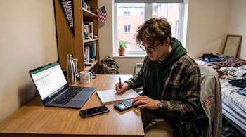 Un joven con gafas escribe en un cuaderno frente a una computadora portátil, un teléfono y una calculadora sobre un escritorio