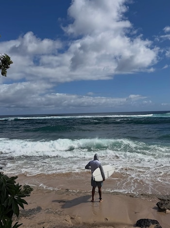 Una persona de espaldas con capucha y bermudas sostiene una tabla de surf en la orilla de una playa de arena frente a olas y mar azul, bajo un cielo con nubes