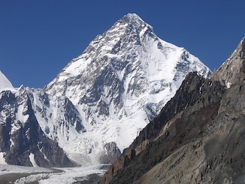 Vista panorámica del pico K2 cubierto de nieve con un glaciar a sus pies y montañas rocosas en primer plano bajo un cielo azul claro