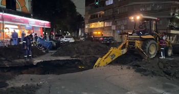 Una excavadora amarilla, un gran socavón en la calle, montones de tierra, vehículos de emergencia, y personal de obra en una calle de noche