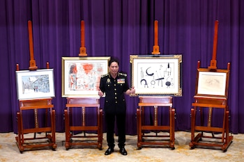 Hombre en uniforme militar negro con medallas, posando entre cuatro caballetes de madera con cuadros, frente a una cortina púrpura