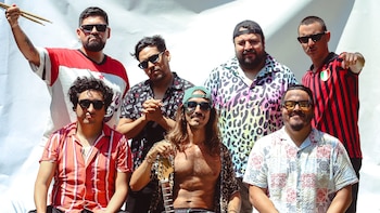 Seis hombres, miembros de una banda, posan para la cámara. Uno sostiene baquetas y otro, en el centro, una guitarra